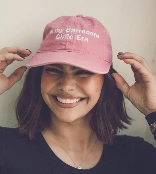 Smiling woman wearing a pink cap embroidered with 'In my Barrecore Girlie Era', celebrating Barrecore lifestyle and merch.