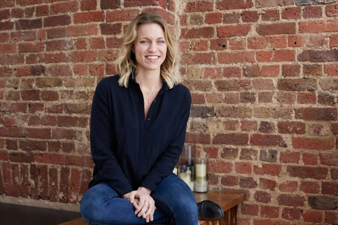 Barrecore founder Niki Rein - a lady with shoulder length blonde hair, wearing blue jeans and a black shirt. Sat on a stool against a bare brick wall.