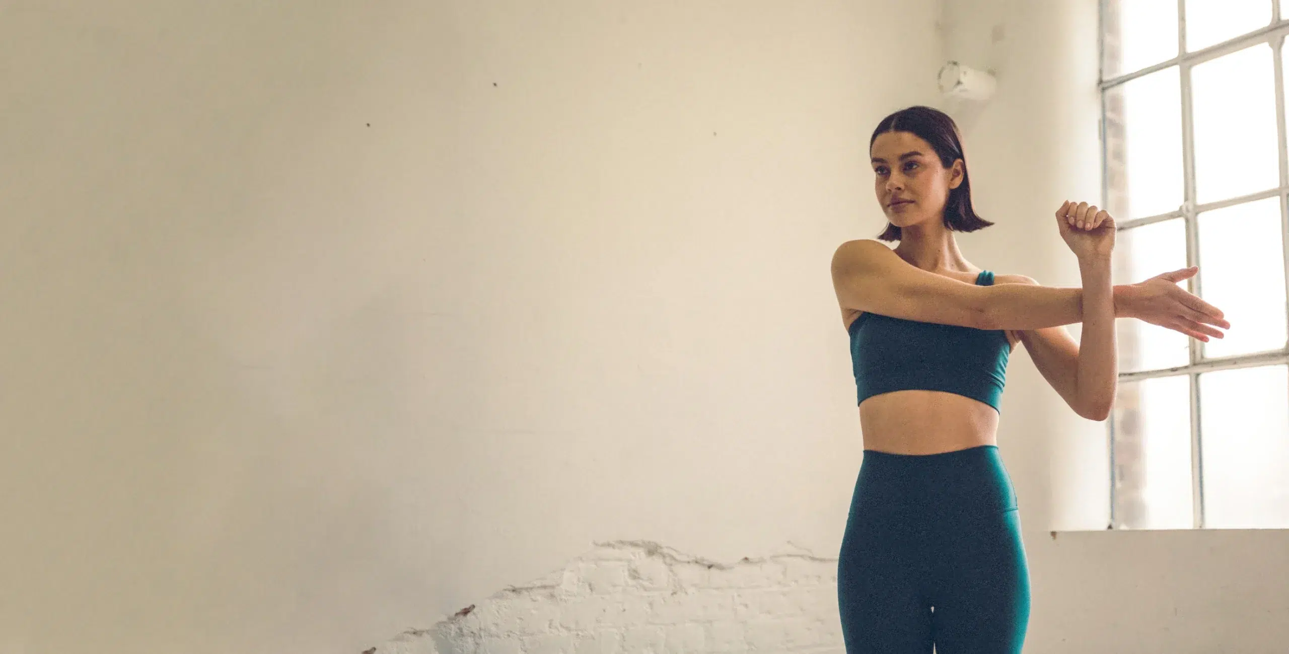 Woman stretching in a Barrecore studio before class, wearing teal activewear in a minimalist, light-filled space.