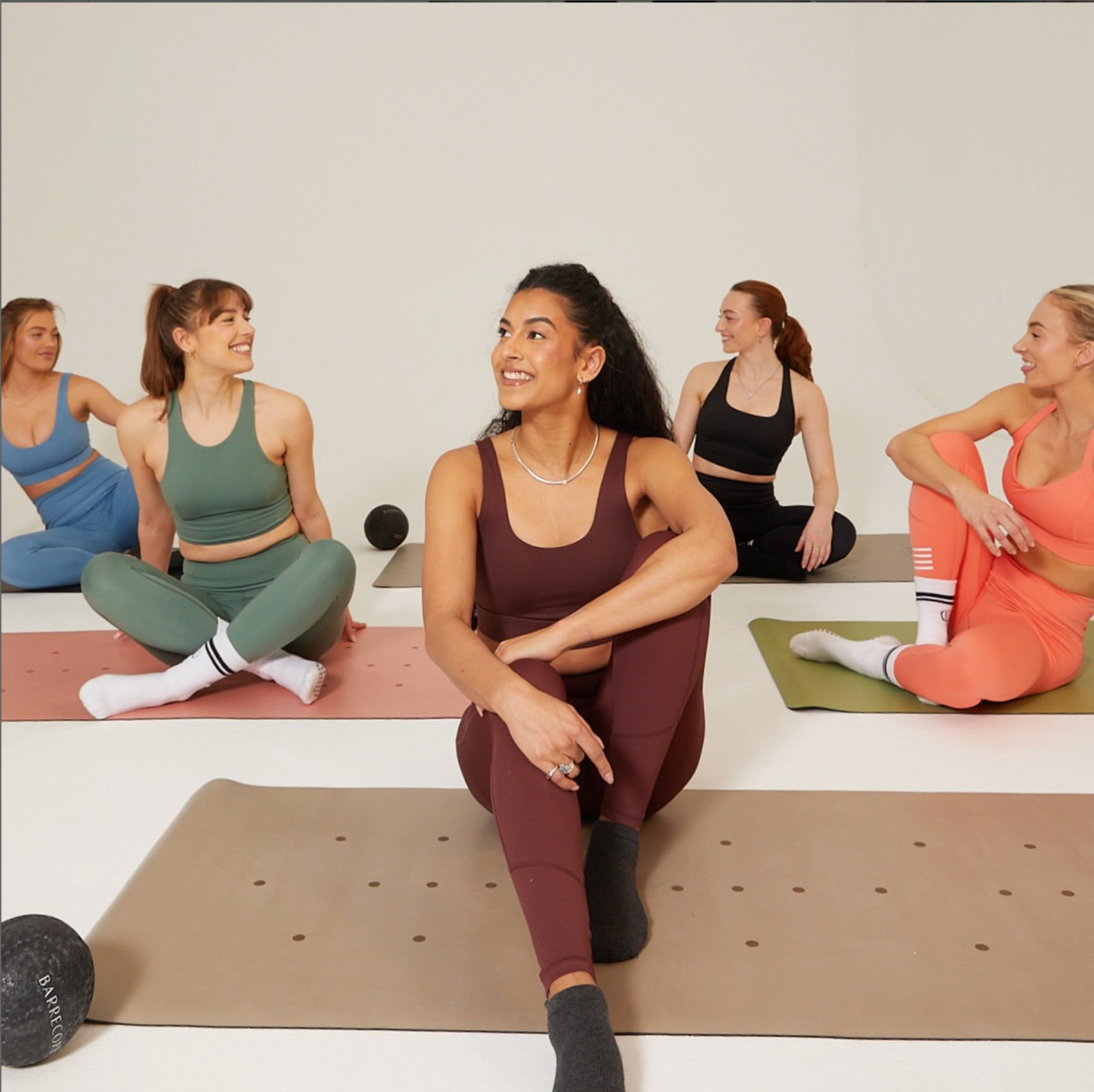 Group of women smiling during a Barrecore class, seated on mats in activewear, mid-stretch in a bright studio.