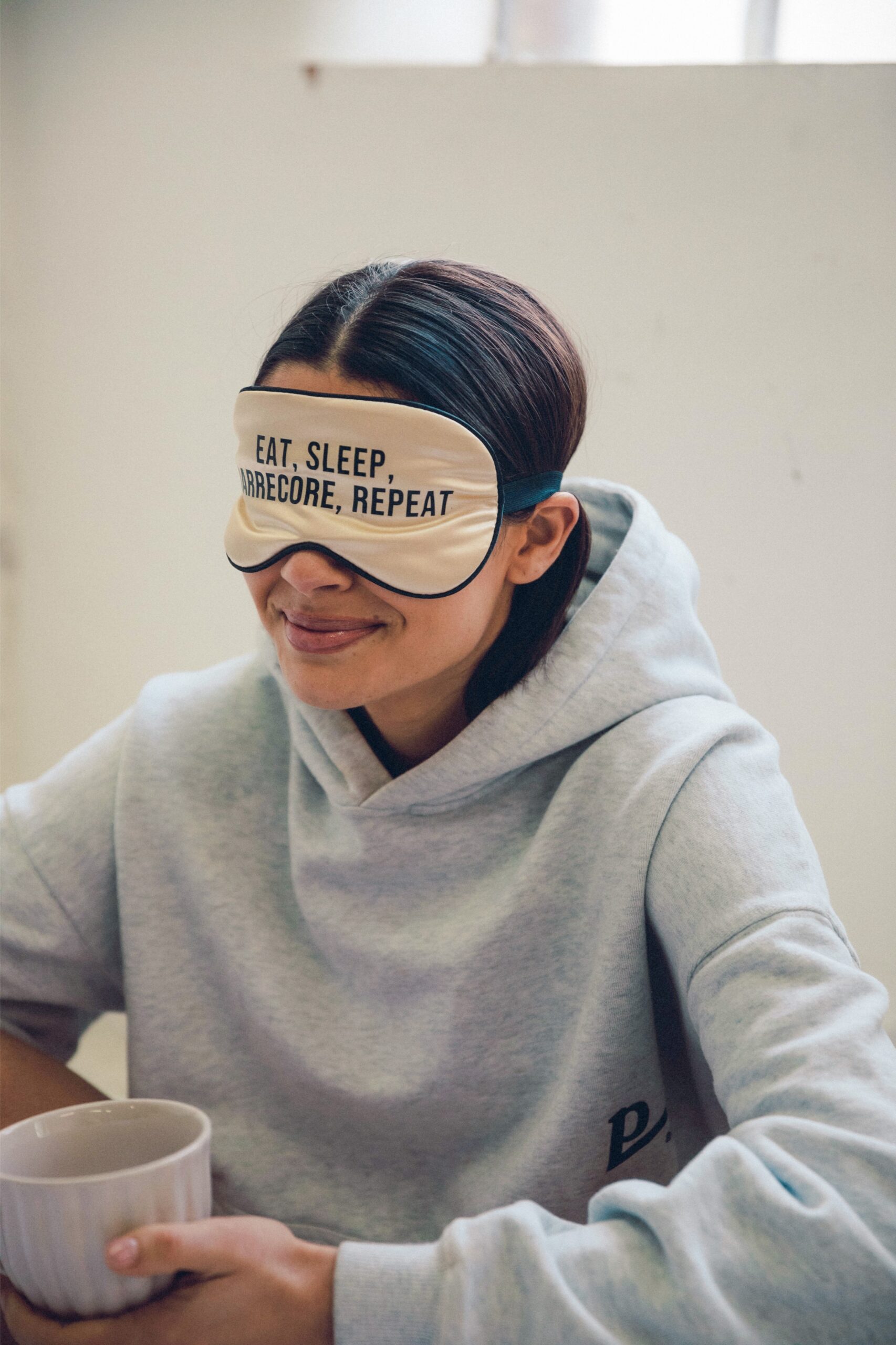 Smiling woman in a hoodie wearing a sleep mask that reads 'Eat, Sleep, Barrecore, Repeat' in a bright studio setting.